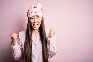 Young asian woman wearing pajama and sleep mask over pink isolated background celebrating surprised and amazed for success with arms raised and open eyes. Winner concept.