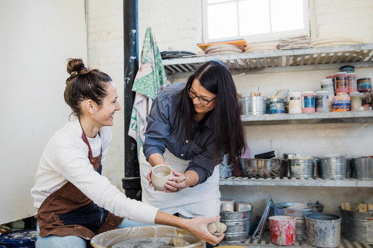 An art teacher giving instruction, help and tips to a student learning to throw clay on a pottery wheel during a lesson. 