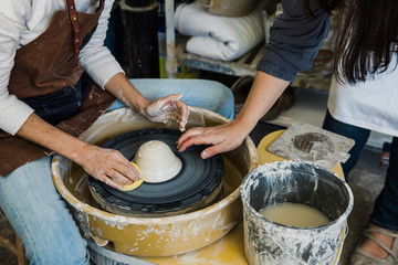 A Caucasian woman with a messy bun leaning over a pottery wheel trying to center her clay with a slip bucket nearby.