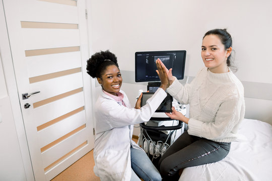 Young African Woman Doctor Consulting And Giving Hi Five With Woman Patient Sitting On The Couch In Modern Hospital. Ultrasound Equipment On The Background