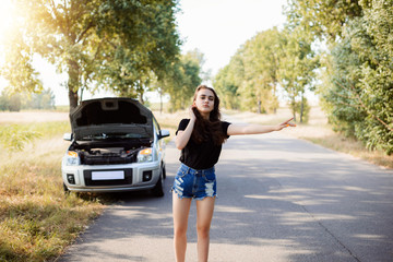 Full length portrait of attractive student girl near modern car. Young girl tries to stop car and ask for a help to start engine of broken car