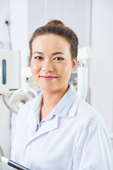 Vertical chest up portrait of modern Asian female dentist wearing white coat looking at camera smiling