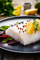 Fresh raw cod with vegetable leaves and lemon  on black plateon on wooden table