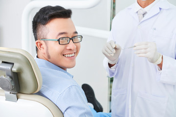 Horizontal shot of happy Asian patient of dental clinic sitting on chair smiling, unrecognizable dentist standing next to him