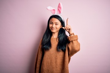 Young beautiful chinese woman wearing bunny ears standing over isolated pink background pointing finger up with successful idea. Exited and happy. Number one.