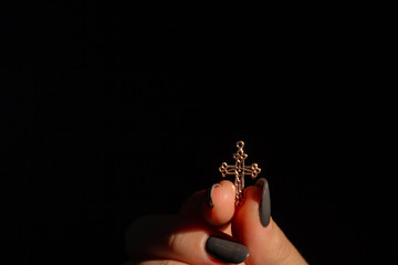 Female's hand is holidng smal golden cross in her fingers. Black background.