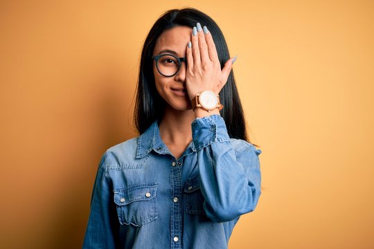 Young beautiful chinese woman wearing casual denim shirt over isolated yellow background covering one eye with hand, confident smile on face and surprise emotion.