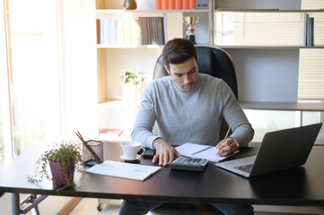 A young man working at a desk with a laptop. Calculator, notepad, pencil, pen and cup of coffee on the side.