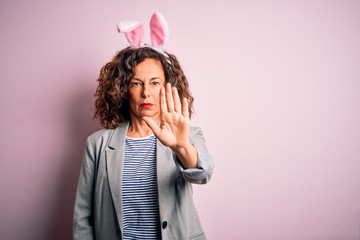 Middle age beautiful woman wearing bunny ears standing over isolated pink background doing stop sing with palm of the hand. Warning expression with negative and serious gesture on the face.
