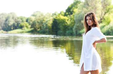 Young sexy brunette woman in white dress standing near river and enjoying sunshine on summer day