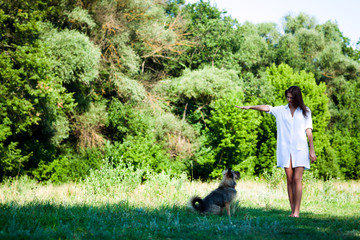 Obraz na płótnie Canvas Young brunette woman in white dress playing with dog on grass on summer day with green trees and river at background