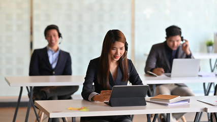 Woman with headset working as call center while working/typing on computer tablet and sitting at her working desk surrounded by her colleagues as background.