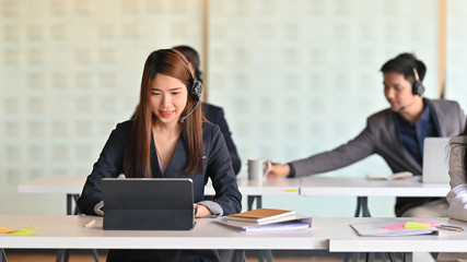 Photo of young beautiful woman with headset working as call center while working/typing on computer tablet and sitting at her working desk surrounded by her colleagues as background.