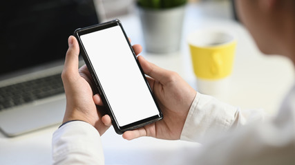 Cropped shot of business man holding on crop black smartphone with white isolated blank screen with the blurred working desk as background.