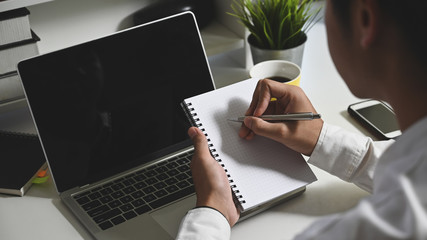 Closeup business man taking notes in front black blank screen laptop at the modern working desk.