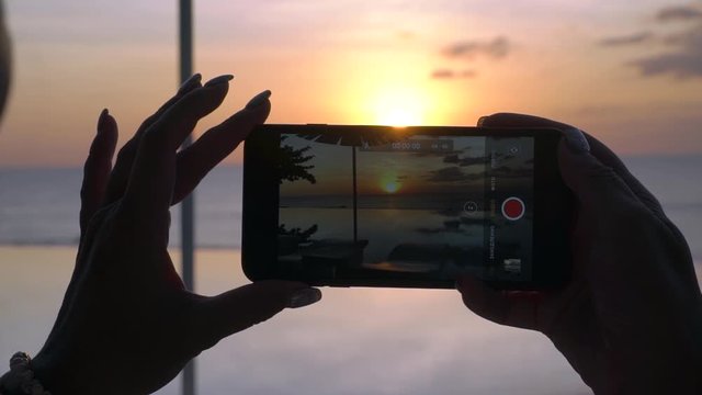 Female hands are holding a phone close-up, and she takes photos and video on the phone a beautiful sunset on a background of the sea. Girl traveler blogger.