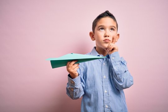 Young little boy kid holding paper plane over isolated pink background serious face thinking about question, very confused idea
