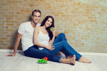 Happy couple with fruits, sitting on floor, close to each other, against loft style wall. Vegetarian, weight lossing, dieting, healthy food concept.