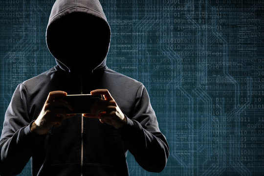 Dangerous hacker with a smartphone gadget over digital background with binary code. Obscured dark face in mask and hood. Data thief, internet attack, darknet fraud, virtual reality and cyber security.