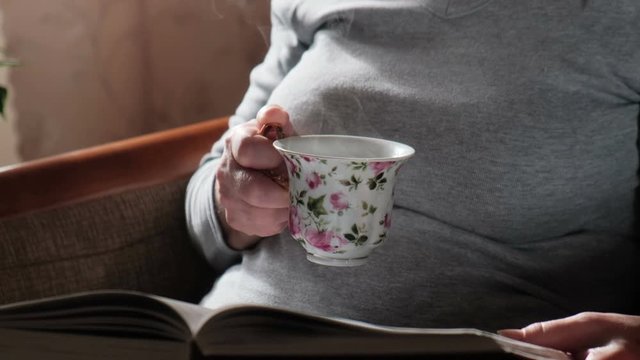 Elderly woman reading a book and drinking hot coffee or tea. Comfort at home