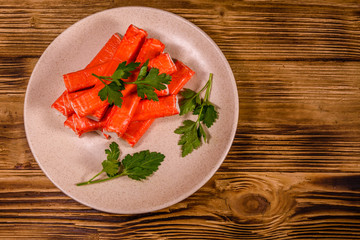 Ceramic plate with pile of crab sticks and parsley twig on a wooden table. Top view