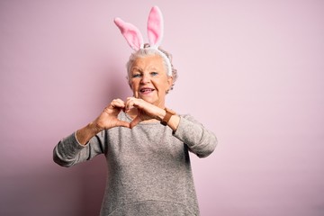 Senior beautiful woman wearing bunny ears standing over isolated pink background smiling in love showing heart symbol and shape with hands. Romantic concept.