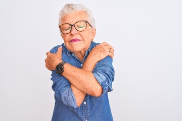 Senior grey-haired woman wearing denim shirt and glasses over isolated white background Hugging oneself happy and positive, smiling confident. Self love and self care