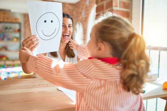Beautiful psycologist and blond toddler girl wearing school uniform sitting on desk doing therapy using emoji emotions at consulting room