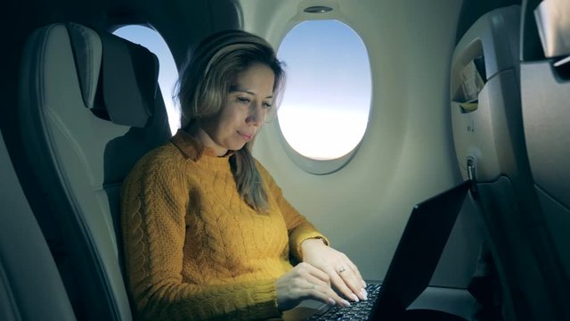 A lady is using a laptop on the plane and smiling. Female passenger working inside of an airplane.