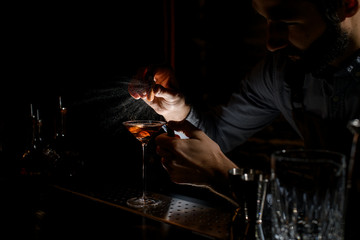 Bartender spraying on the alcoholic drink in a martini glass with orage zest juice in the dark