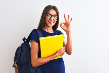 Young student woman wearing backpack glasses holding book over isolated white background doing ok sign with fingers, excellent symbol