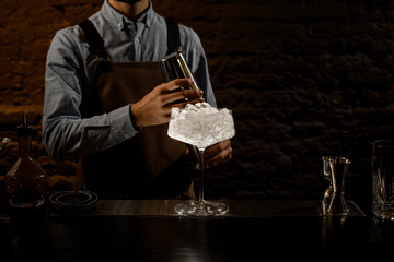 Bartender holding a steel shaker on the bar counter with the big cocktail glass full of ice
