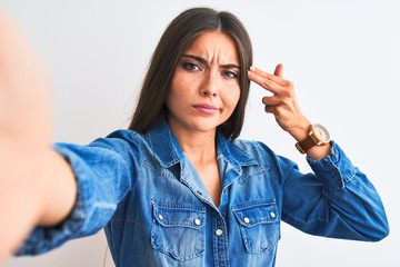 Beautiful woman wearing denim shirt make selfie by camera over isolated white background Shooting and killing oneself pointing hand and fingers to head like gun, suicide gesture.