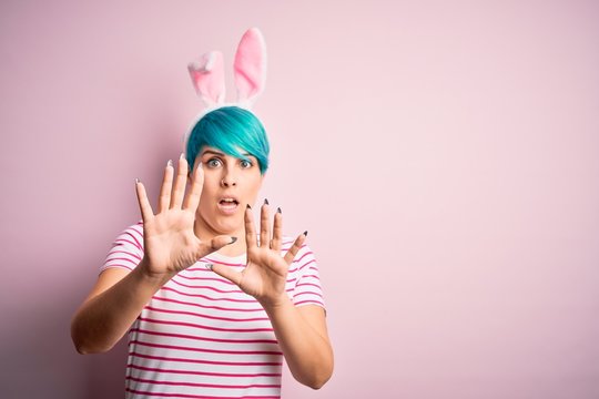Young woman with fashion blue hair wearing easter rabbit ears over pink background afraid and terrified with fear expression stop gesture with hands, shouting in shock. Panic concept.