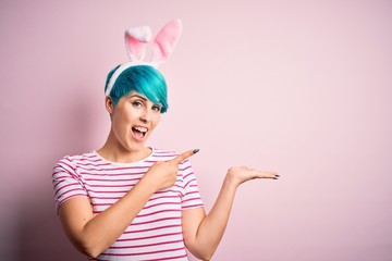 Young woman with fashion blue hair wearing easter rabbit ears over pink background amazed and smiling to the camera while presenting with hand and pointing with finger.