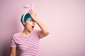 Young woman with fashion blue hair wearing easter rabbit ears over pink background surprised with hand on head for mistake, remember error. Forgot, bad memory concept.