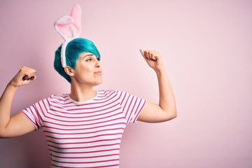 Young woman with fashion blue hair wearing easter rabbit ears over pink background showing arms...