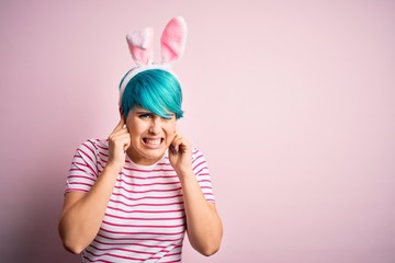 Young woman with fashion blue hair wearing easter rabbit ears over pink background covering ears...