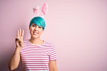 Young woman with fashion blue hair wearing easter rabbit ears over pink background showing and pointing up with fingers number three while smiling confident and happy.