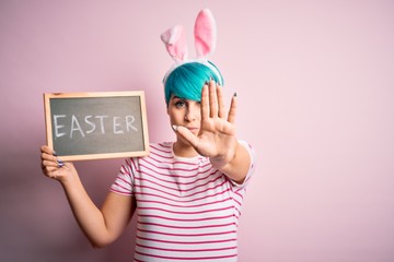Young woman with fashion blue hair wearing easter rabbit ears and holding blackboard with holiday word with open hand doing stop sign with serious and confident expression, defense gesture