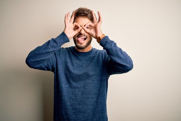 Young handsome man with beard wearing casual sweater standing over white background doing ok gesture like binoculars sticking tongue out, eyes looking through fingers. Crazy expression.