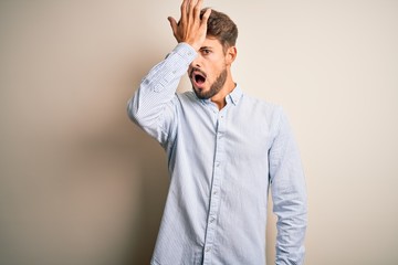 Young handsome man with beard wearing striped shirt standing over white background surprised with...