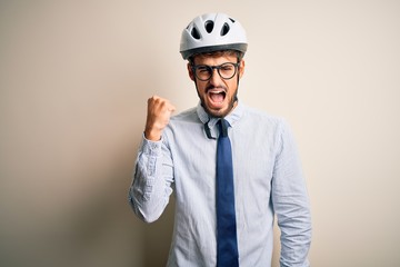 Young businessman wearing glasses and bike helmet standing over isolated white bakground angry and mad raising fist frustrated and furious while shouting with anger. Rage and aggressive concept.