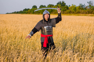 Naklejka na ściany i meble A medieval nomad warrior in oriental armor on the background of a wheat field. Standing in a fencing rack.