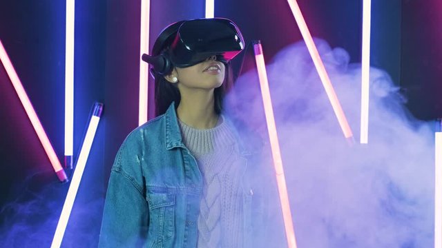 Young woman looks around and wonders how amazing in the virtual reality. Virtual reality helmet on color lighting smoke background
