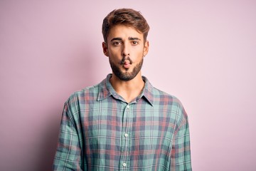 Young handsome man with beard wearing casual shirt standing over pink background making fish face with lips, crazy and comical gesture. Funny expression.
