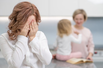 A teenage girl is crying, in the background her mother is playing with her younger sister. Problems of puberty, depression, problems in the family