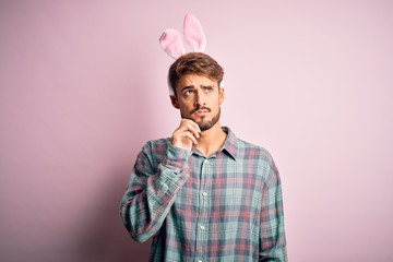 Young handsome adult man wearing cute easter rabbit ears over pink isolated background with hand on chin thinking about question, pensive expression. Smiling with thoughtful face. Doubt concept.