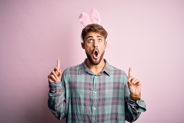 Young handsome adult man wearing cute easter rabbit ears over pink isolated background amazed and surprised looking up and pointing with fingers and raised arms.