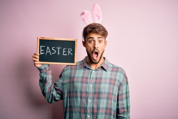 Young handsome man wearing cute easter rabbit ears and holding blackboard with holiday word scared in shock with a surprise face, afraid and excited with fear expression